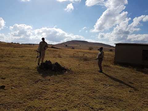 Two people in field gear standing in a wide, grassy field with a building and hill in the background.